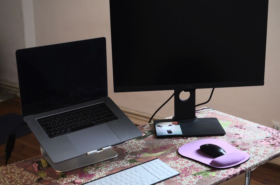 A laptop raised on a stand next to an external monitor, keyboard, and mouse on a desk